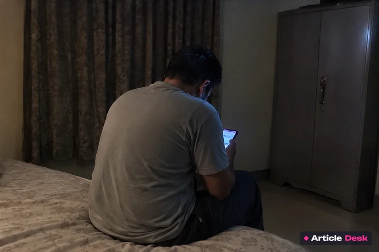 A worried man sitting on the edge of his bed, reading a message on his phone